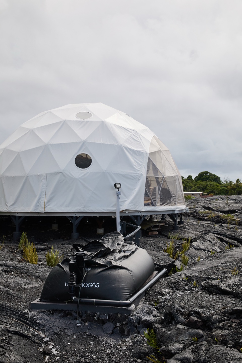A black double-membrane biogas holder sitting on a plinth in the lava field, piped to a white geodesic dome raised on posts behind it.