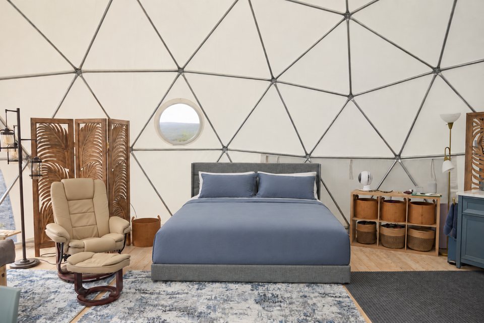 A dome bedroom at The Albizia: a grey upholstered bed with blue linen, a cream leather recliner and a carved wooden screen beneath the white geodesic frame, a round window above.