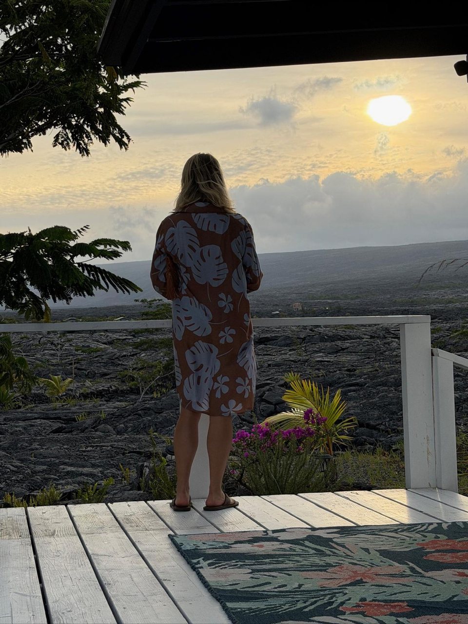 A guest in a leaf-print dress standing at the rail of a white deck, looking out over the lava field toward the setting sun.