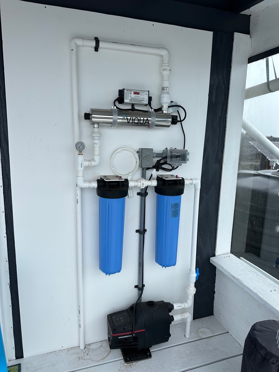 The water plant on the property: two blue cartridge filter housings, a stainless ultraviolet steriliser, a pressure gauge and a pump, plumbed in white PVC against a white wall.